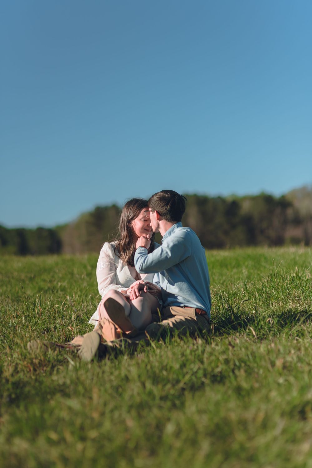 Couple sitting on green grass field sharing a romantic moment under a clear blue sky on a sunny day.