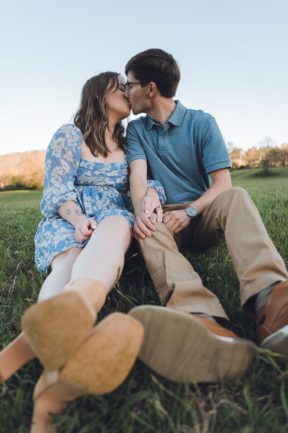 Couple kissing while sitting on green grass outdoors, woman in blue floral dress holding hands with man in blue polo shirt.