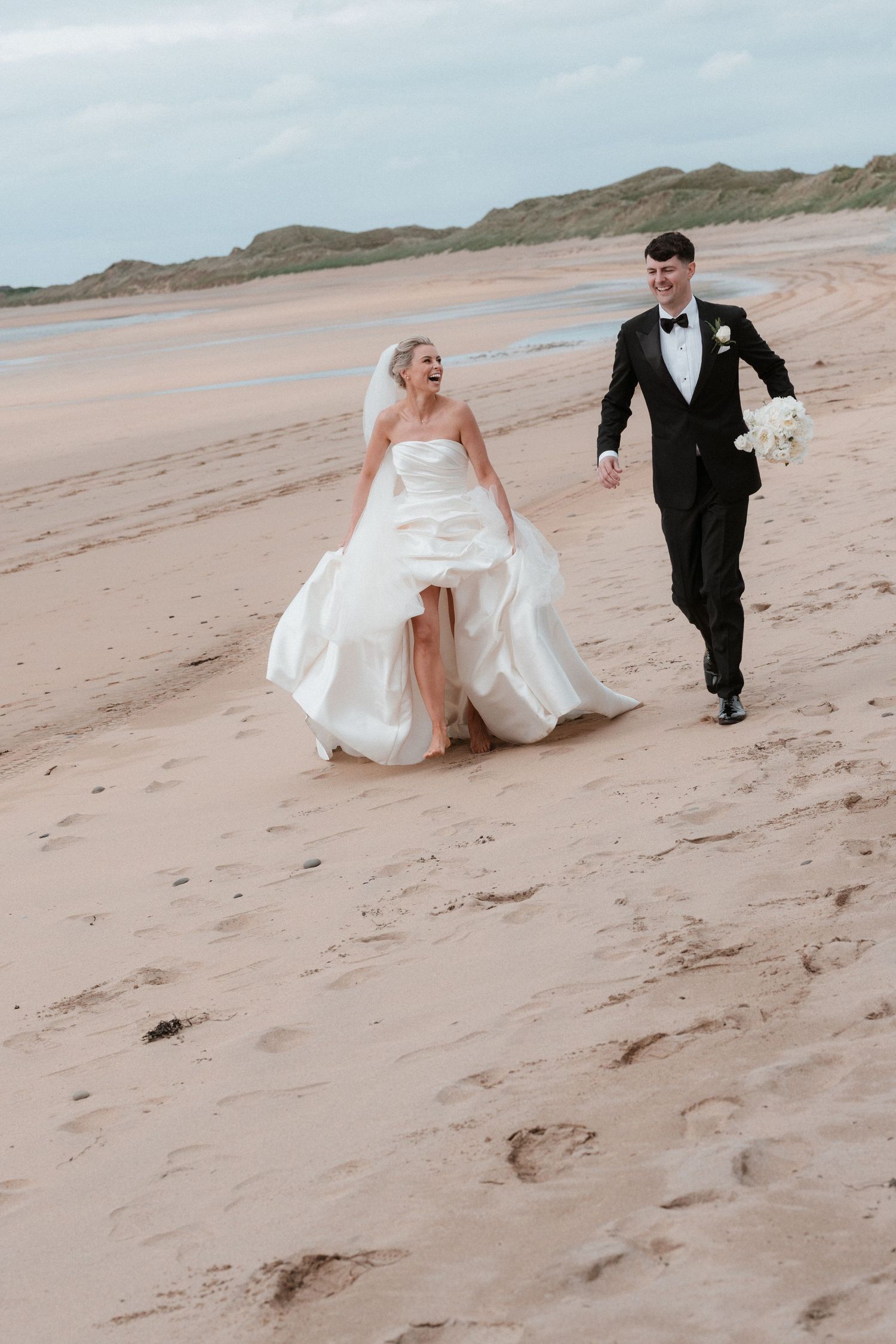 Bride and groom walking hand in hand on a sandy beach, bride in white gown with veil, groom in black tuxedo.