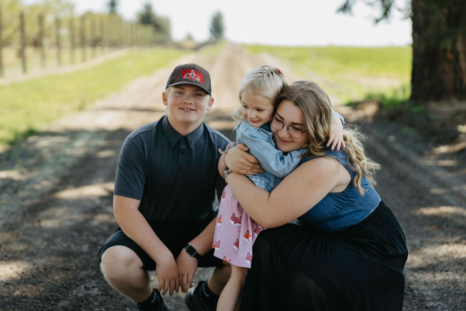 Family portrait of a boy, young girl, and woman hugging on a rural dirt path surrounded by trees.