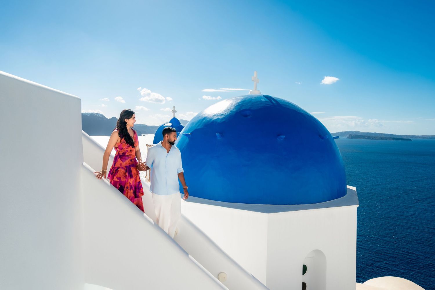Couple walking near iconic blue-domed church in Santorini, Greece, with stunning Aegean Sea views.
