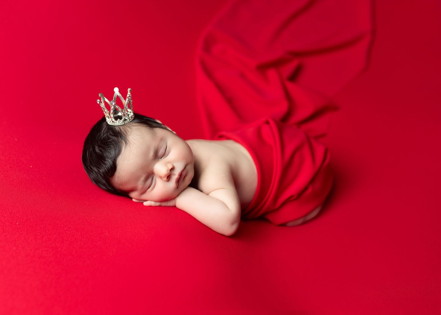 Sleeping newborn baby wearing a tiny crown on a red background, wrapped in red fabric.