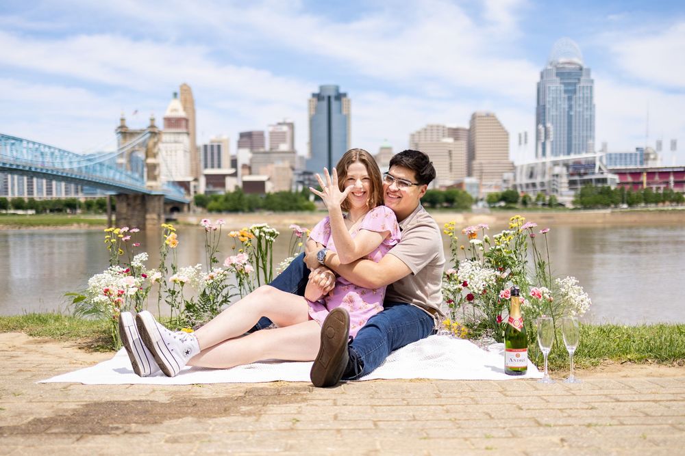 Couple sitting on white blanket by Cincinnati riverfront with city skyline and floral decor during outdoor engagement session.