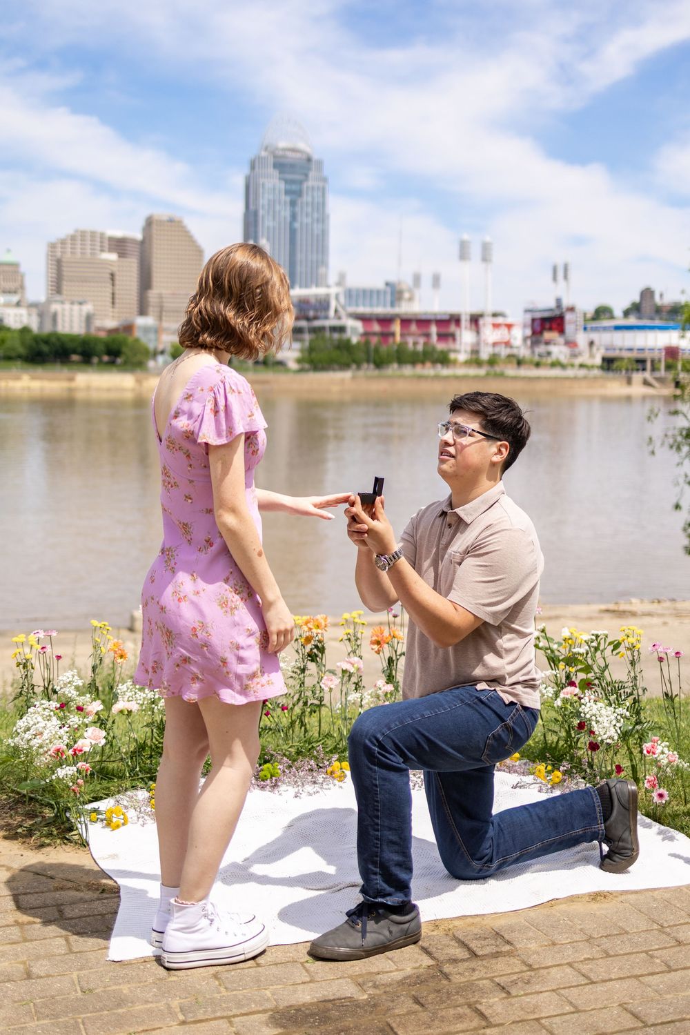 Man proposes on one knee to woman in pink dress by Ohio river with Cincinnati skyline backdrop on sunny summer day.