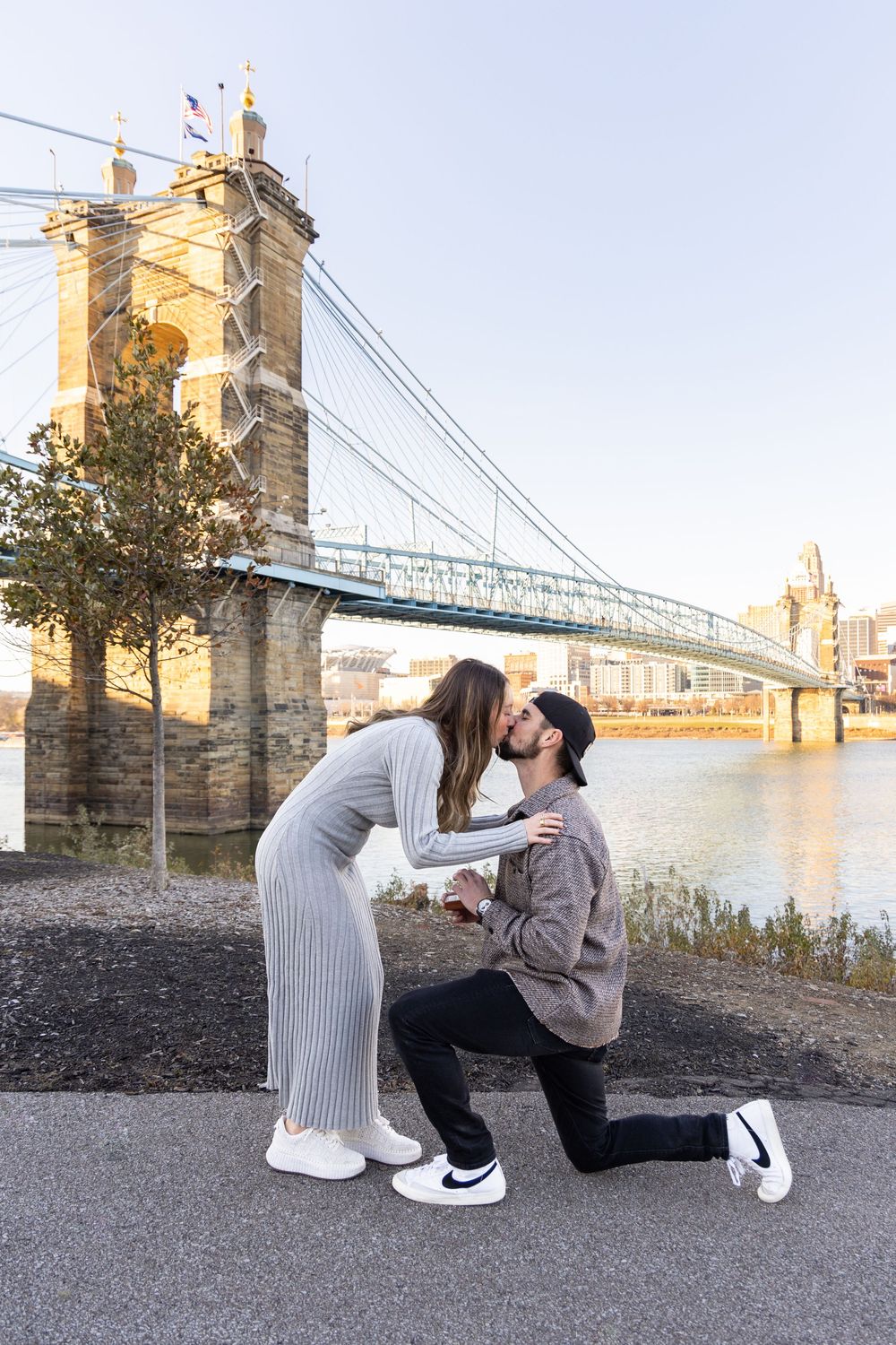 Couple sharing a kiss during a marriage proposal by the John A. Roebling Suspension Bridge in Cincinnati.