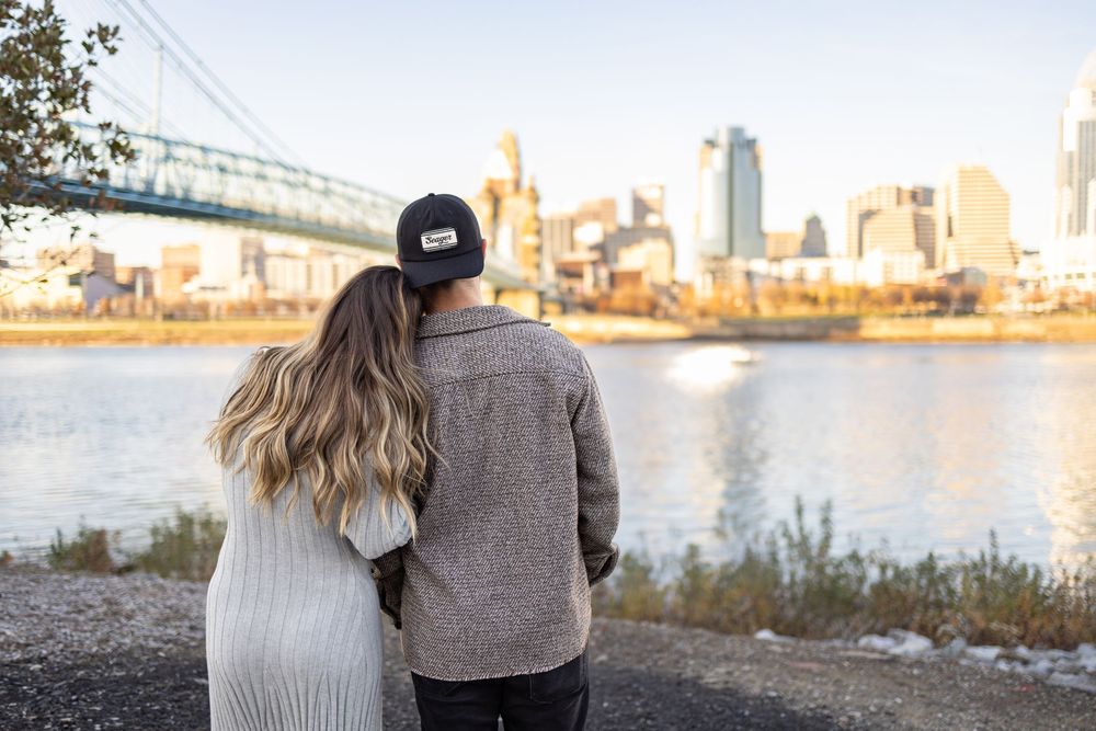Couple embracing while overlooking the Ohio river with Cincinnati skyline and Roebling suspension bridge in the background.