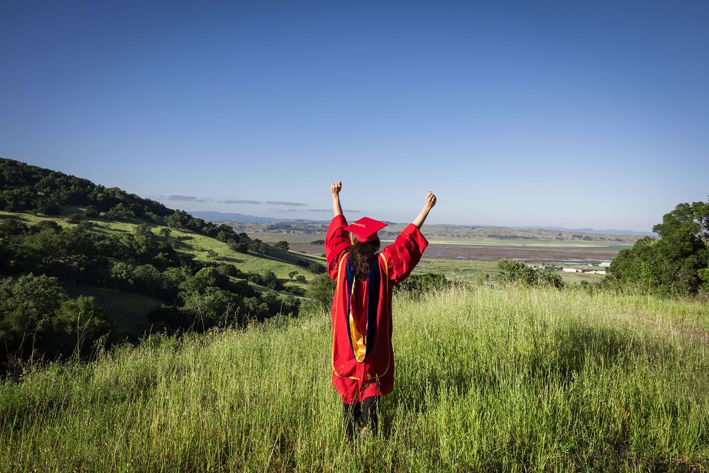 Graduate in red cap and gown stands on green hillside with arms raised in celebration against clear blue sky.