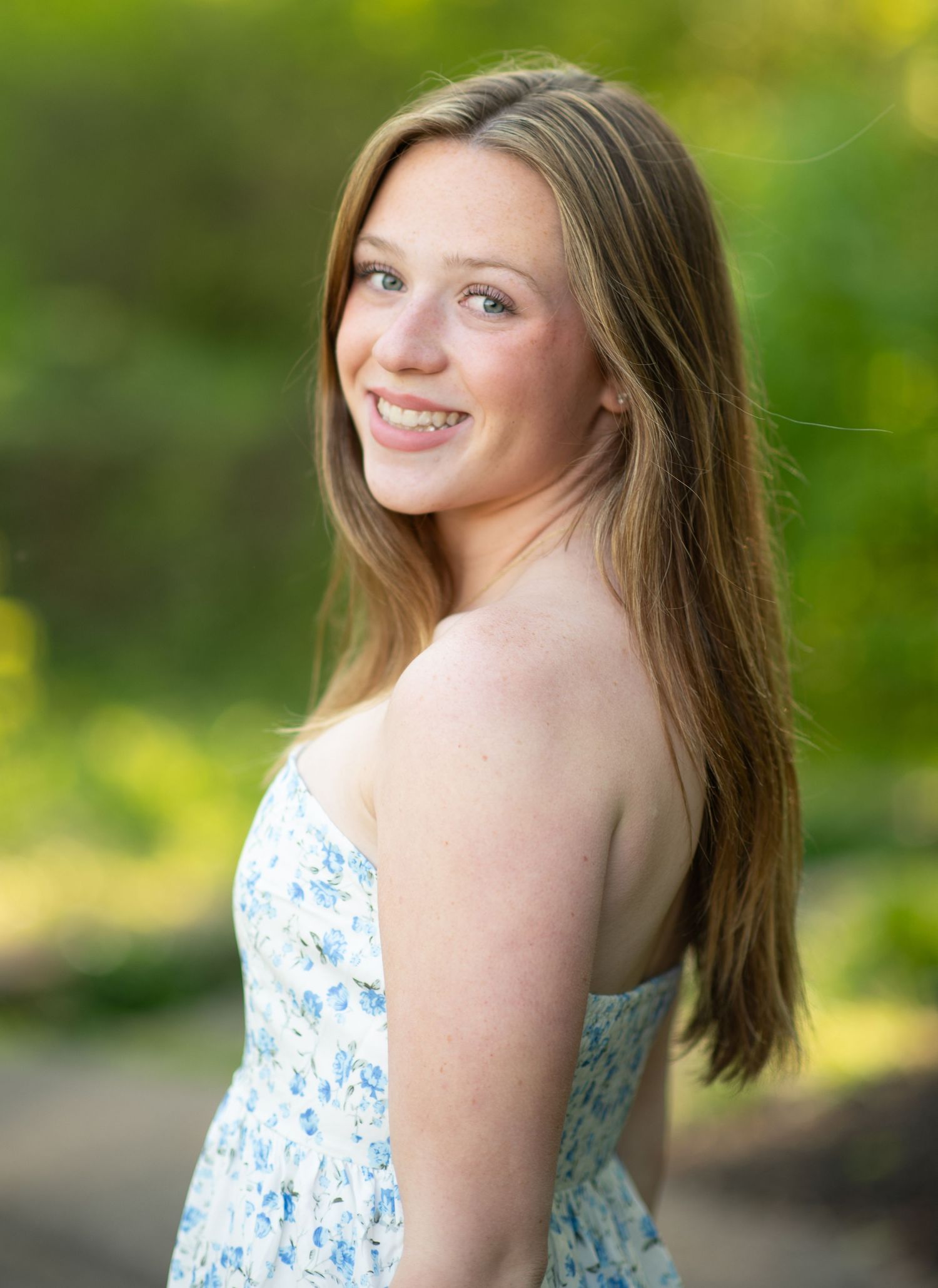 Young woman in floral sundress smiling over shoulder outdoors in lush green garden setting.