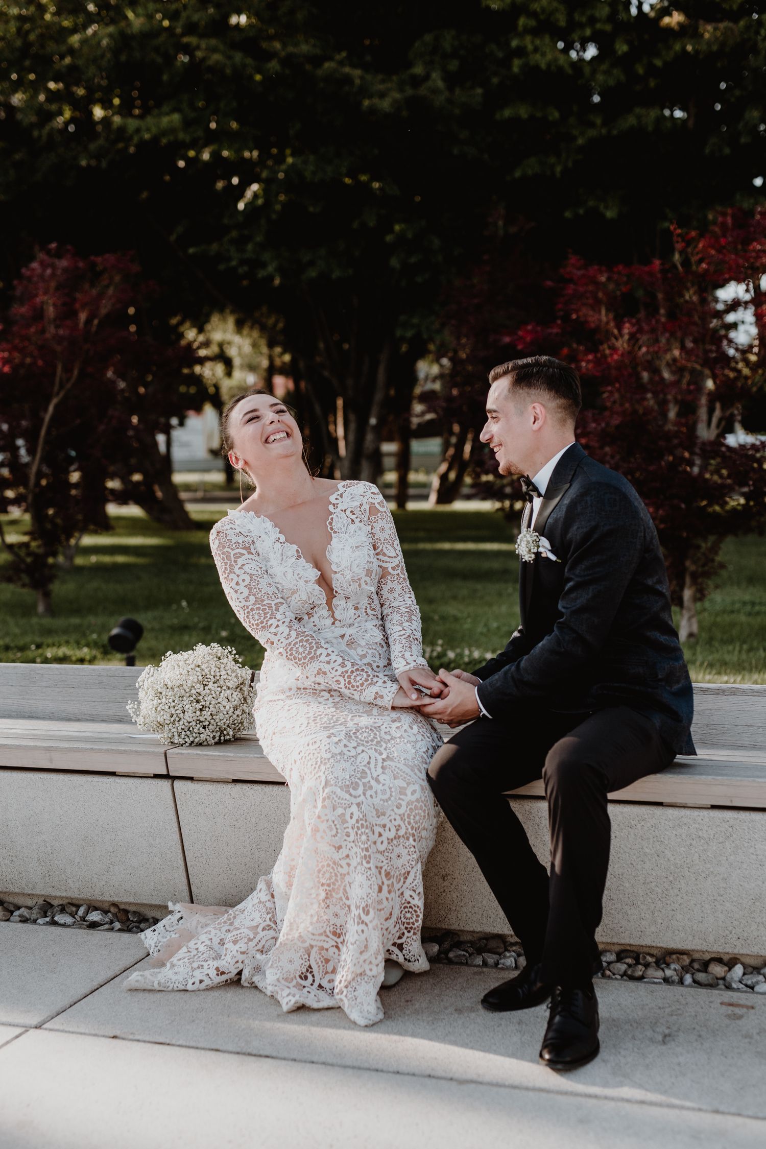 A joyful couple sits together on a concrete bench, the bride wearing an intricate lace gown and the groom in a classic tuxedo.