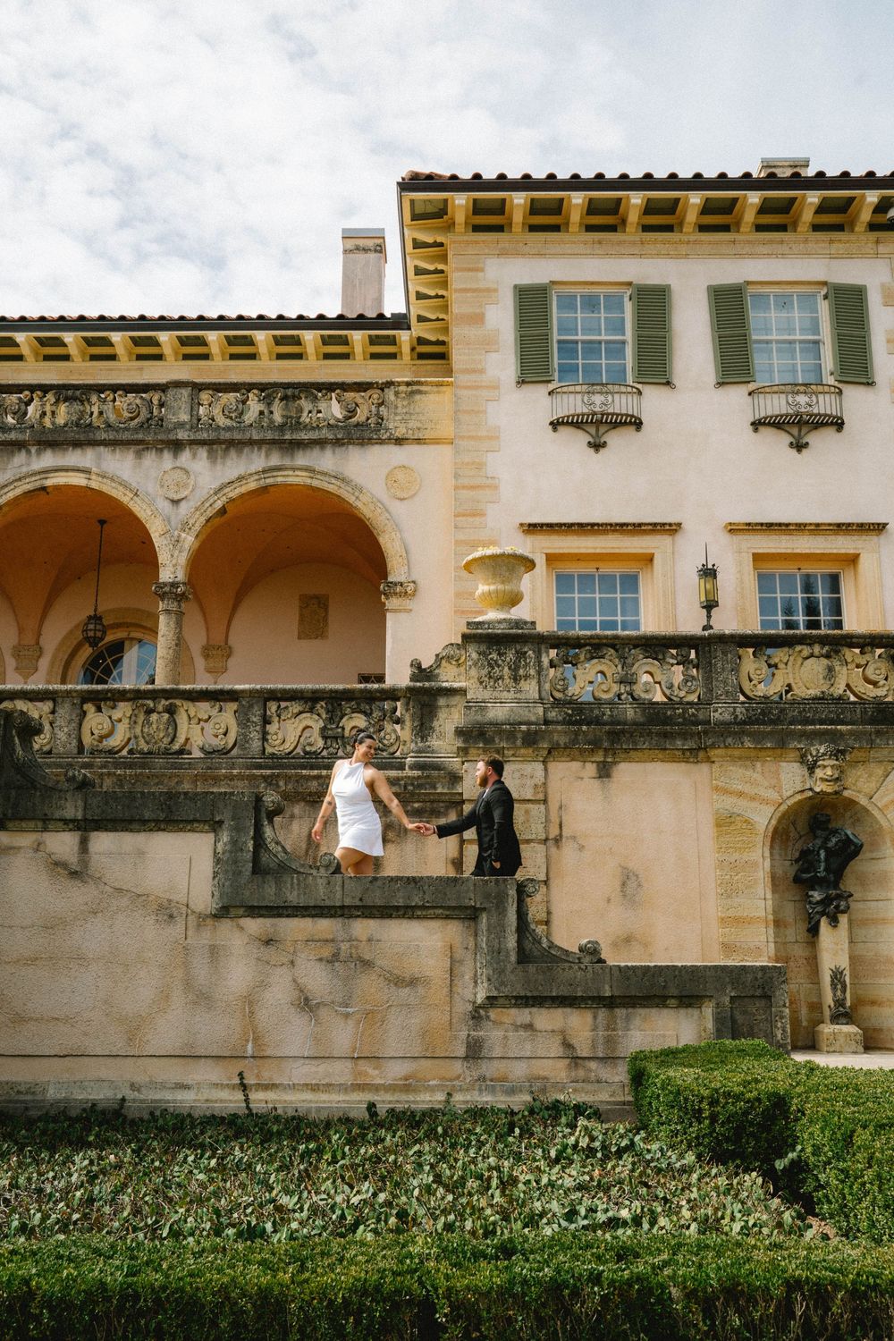 Couple holding hands on ornate stone steps of elegant Mediterranean villa with arched colonnade and lush hedges.