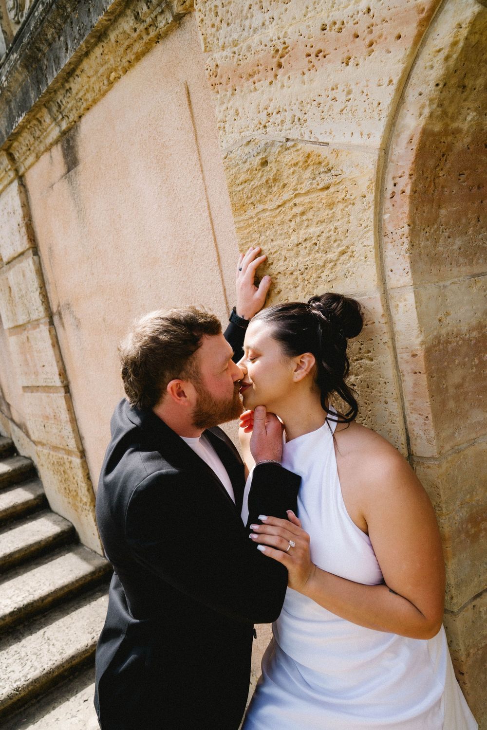 Bride and groom share a romantic almost-kiss against a stone wall, she in a white halter gown, he in a black suit.
