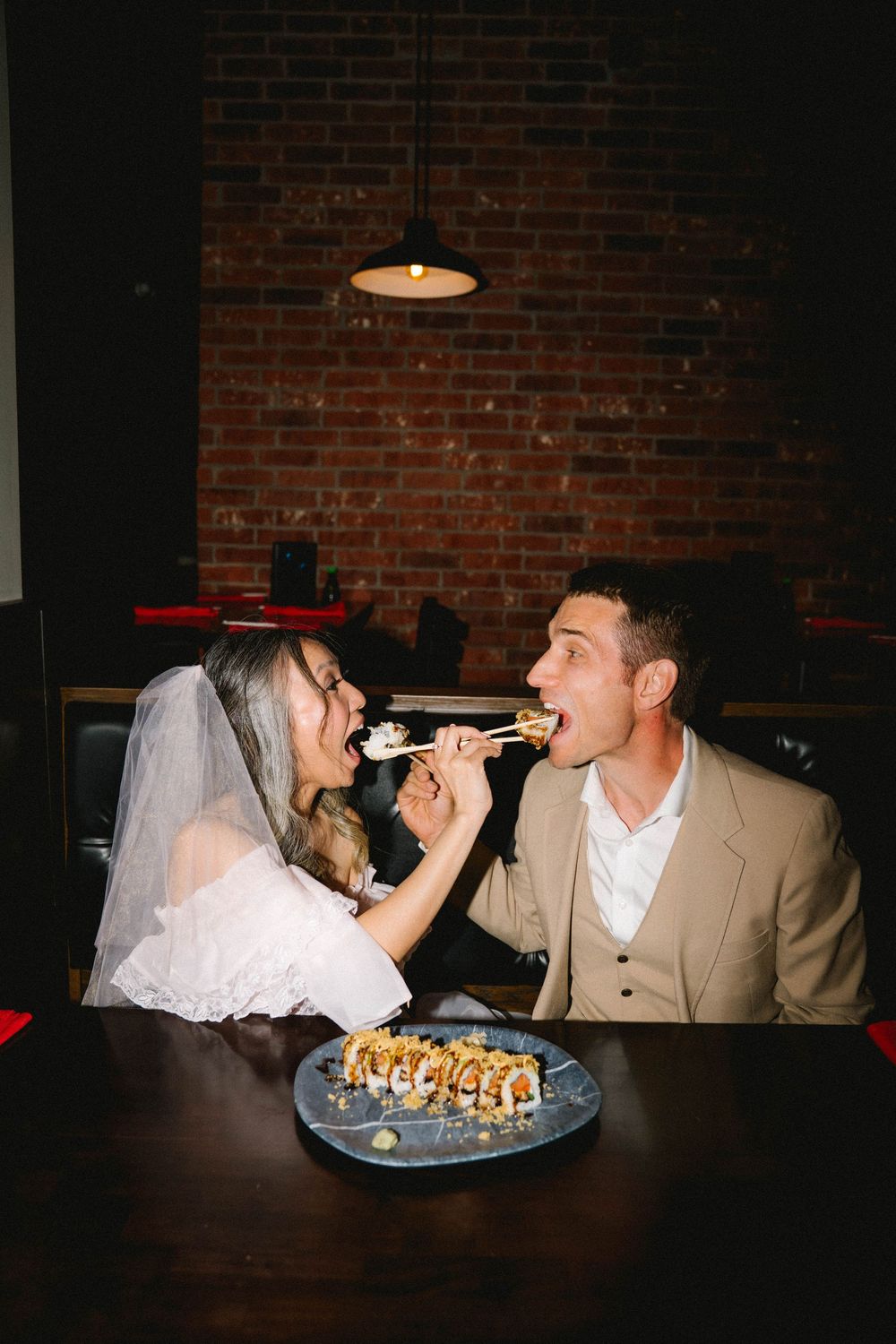Bride and groom share sushi with chopsticks at a romantic restaurant with exposed brick walls.
