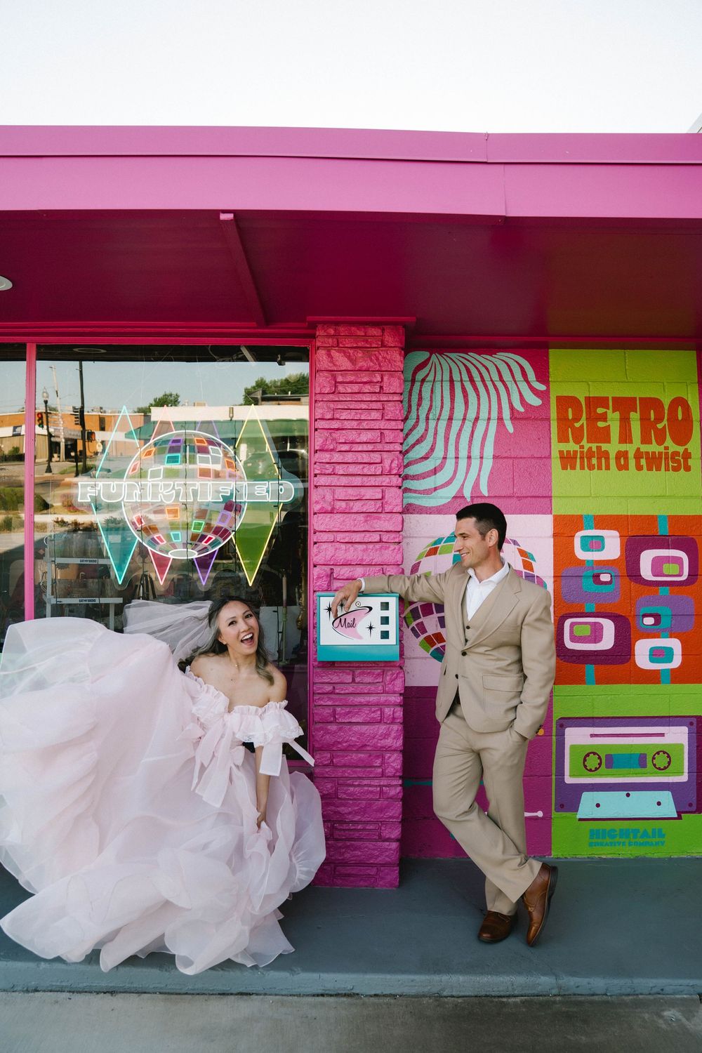 Bride in pink gown and groom in tan suit pose playfully outside a colorful retro-themed building.