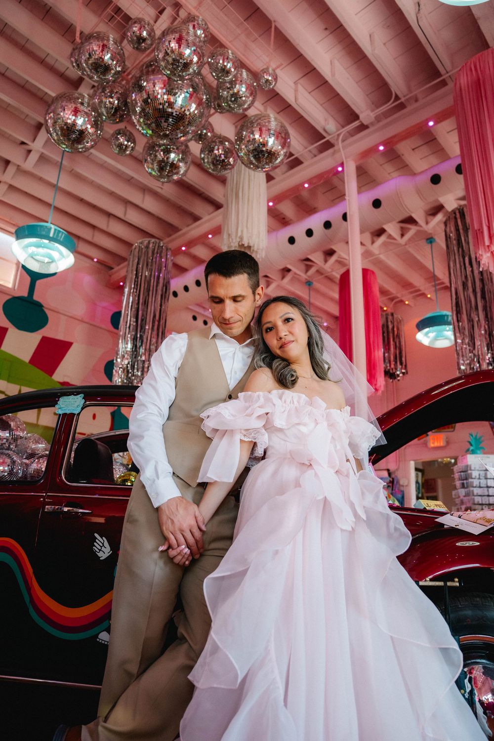 Elegant bride in pink ruffled gown leans on groom in tan vest inside colorful venue with disco ball ceiling.