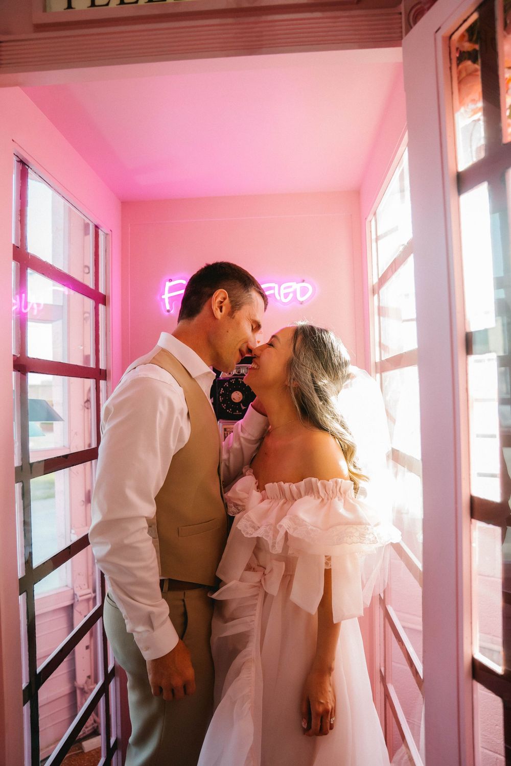 Bride and groom sharing a romantic kiss in a pink neon-lit room, bride wearing an off-shoulder white dress.