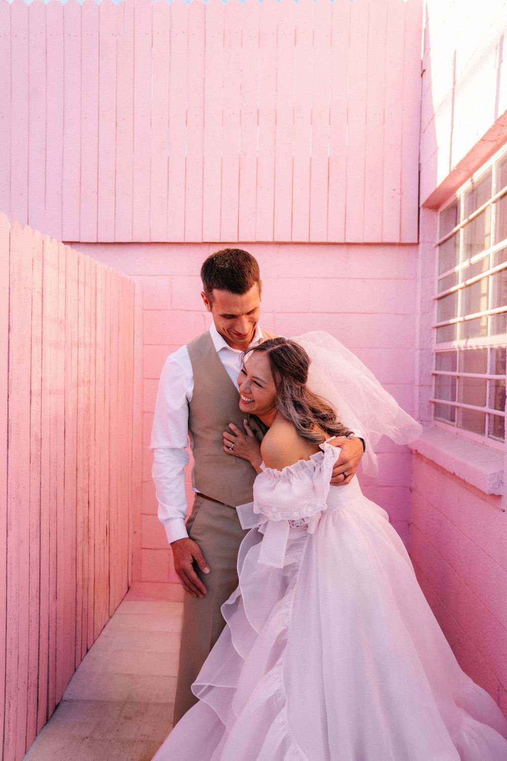 Bride and groom embrace tenderly in a narrow pink-walled corridor, her veil flowing in the breeze.