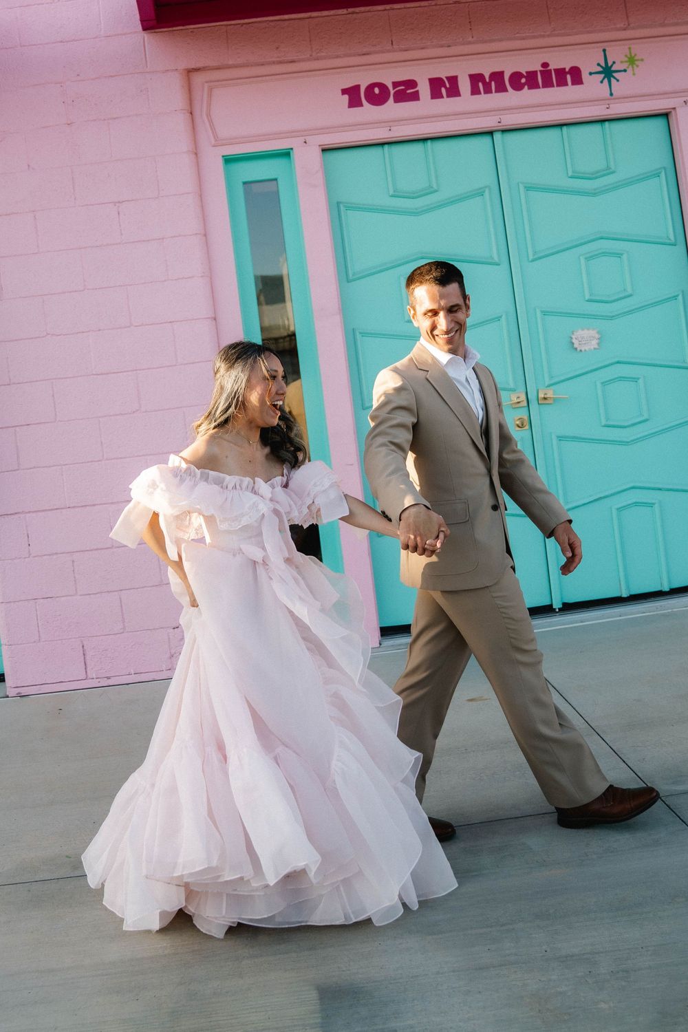 Bride in white ruffled gown and groom in tan suit hold hands walking outside a pink building at 102 N Main.