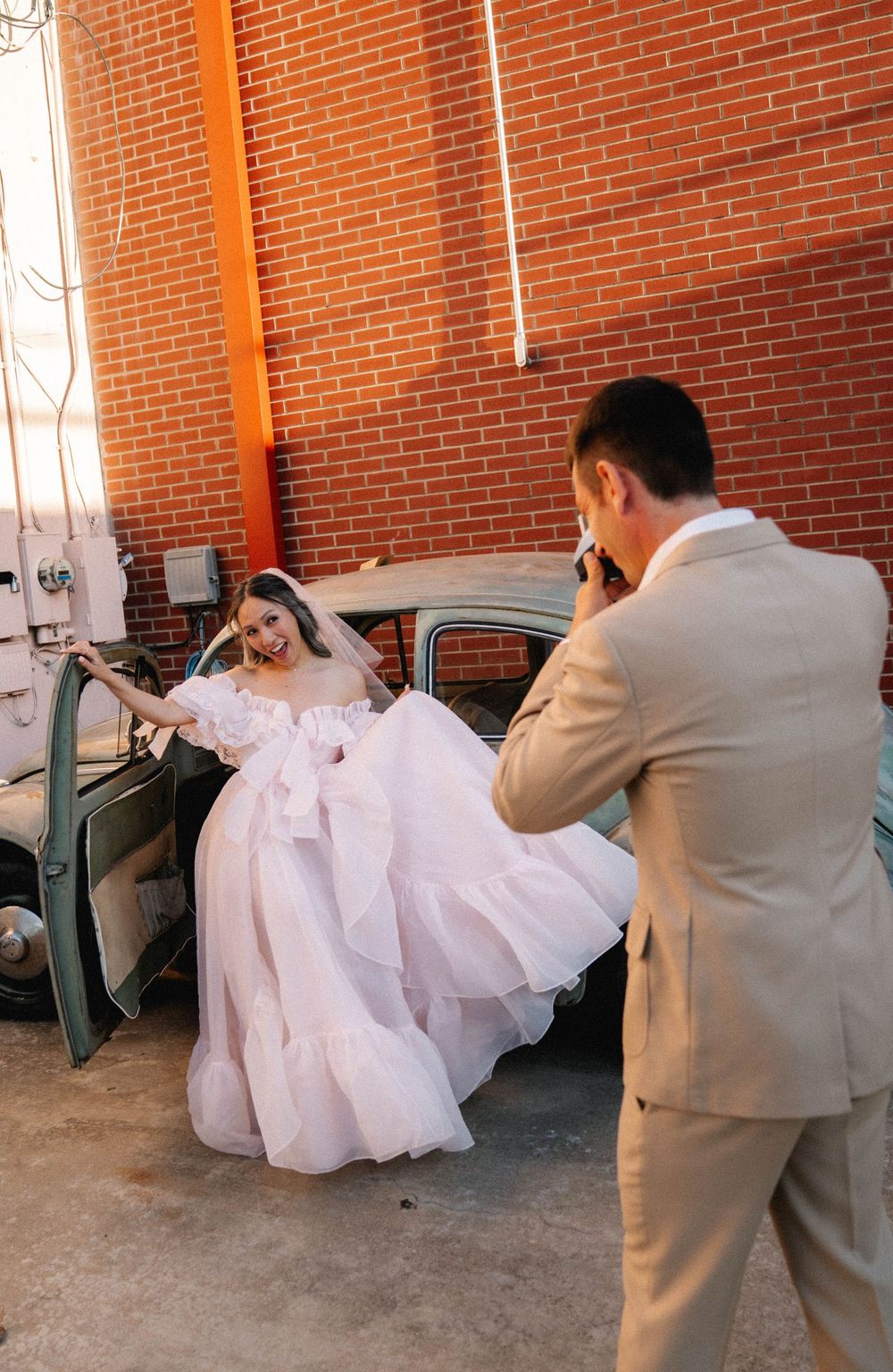 Groom in tan suit photographs bride in white ball gown posing by vintage car against brick wall.