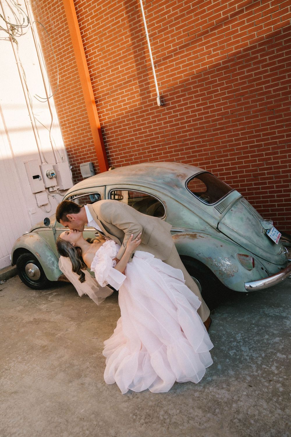 Bride and groom share a romantic dip kiss in front of a vintage teal VW Beetle against a rustic brick wall.