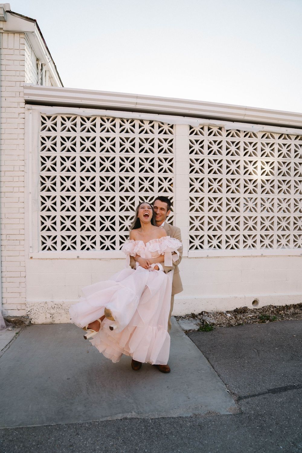 A joyful couple in wedding attire dances outdoors, the bride in a flowing pink off-shoulder gown, against a white lattice wall.