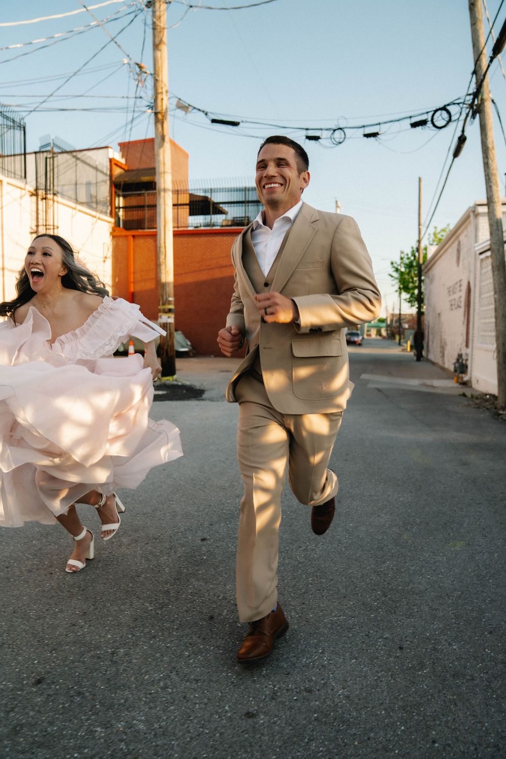 Joyful bride and groom running together in an alley, bride in pink gown, groom in tan suit, on their wedding day.