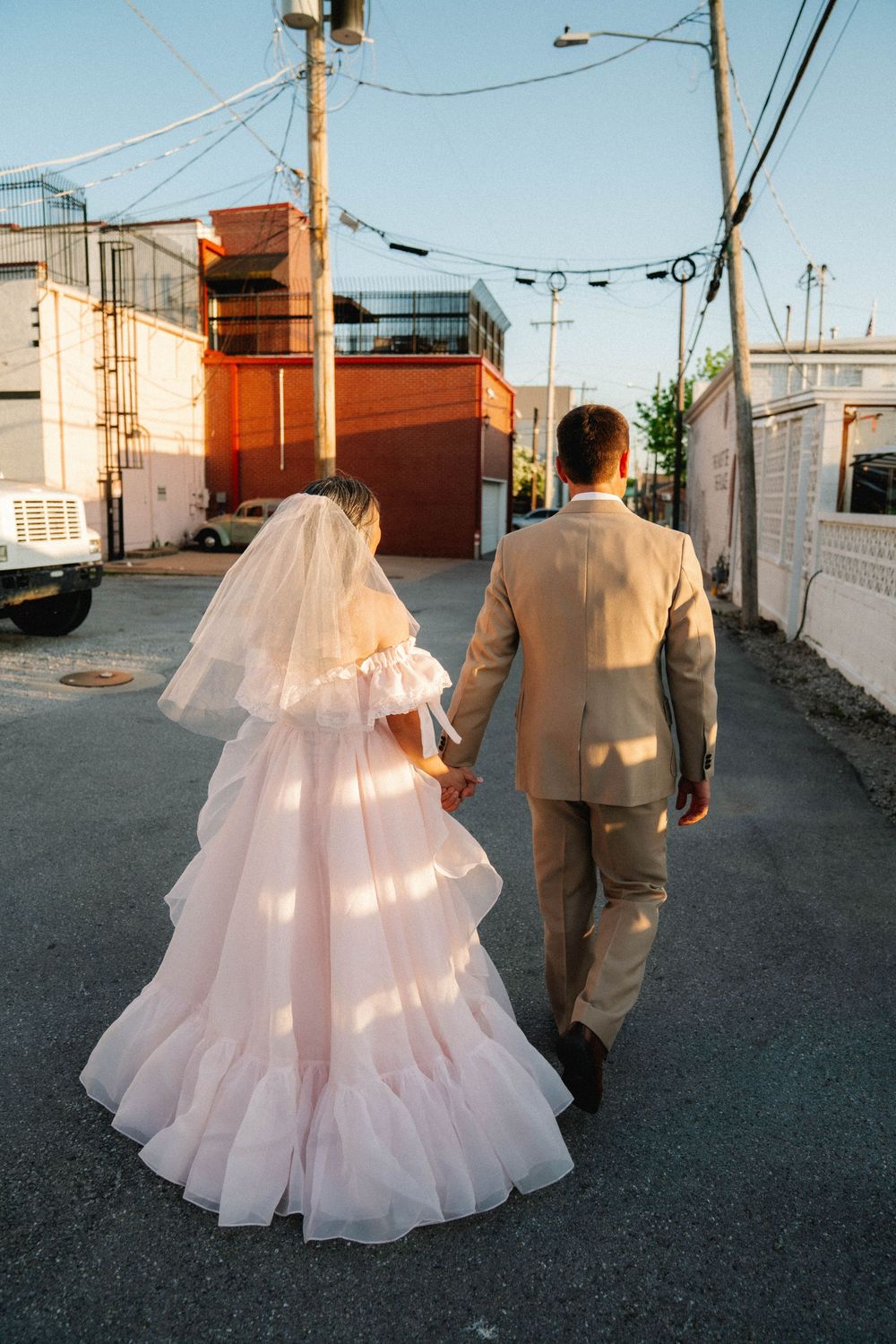 Bride in pink ruffled gown with veil and groom in tan suit walk hand in hand down an urban alley at sunset.