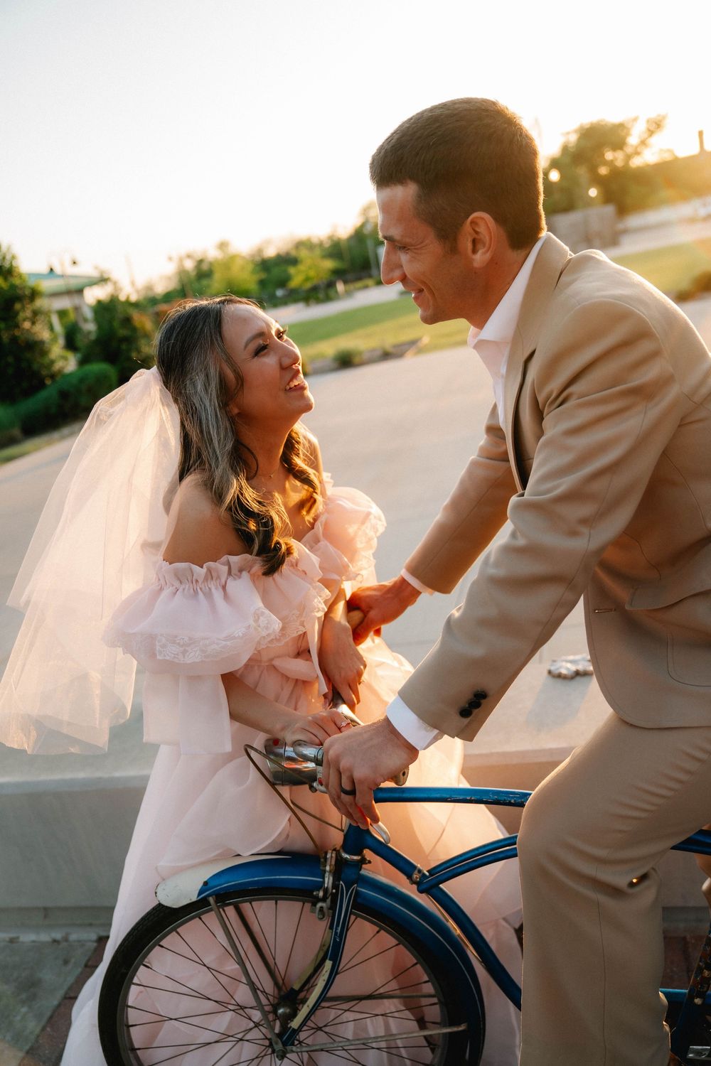 Bride in white dress and veil sits on blue bicycle with groom in tan suit during golden hour outdoor wedding photo.