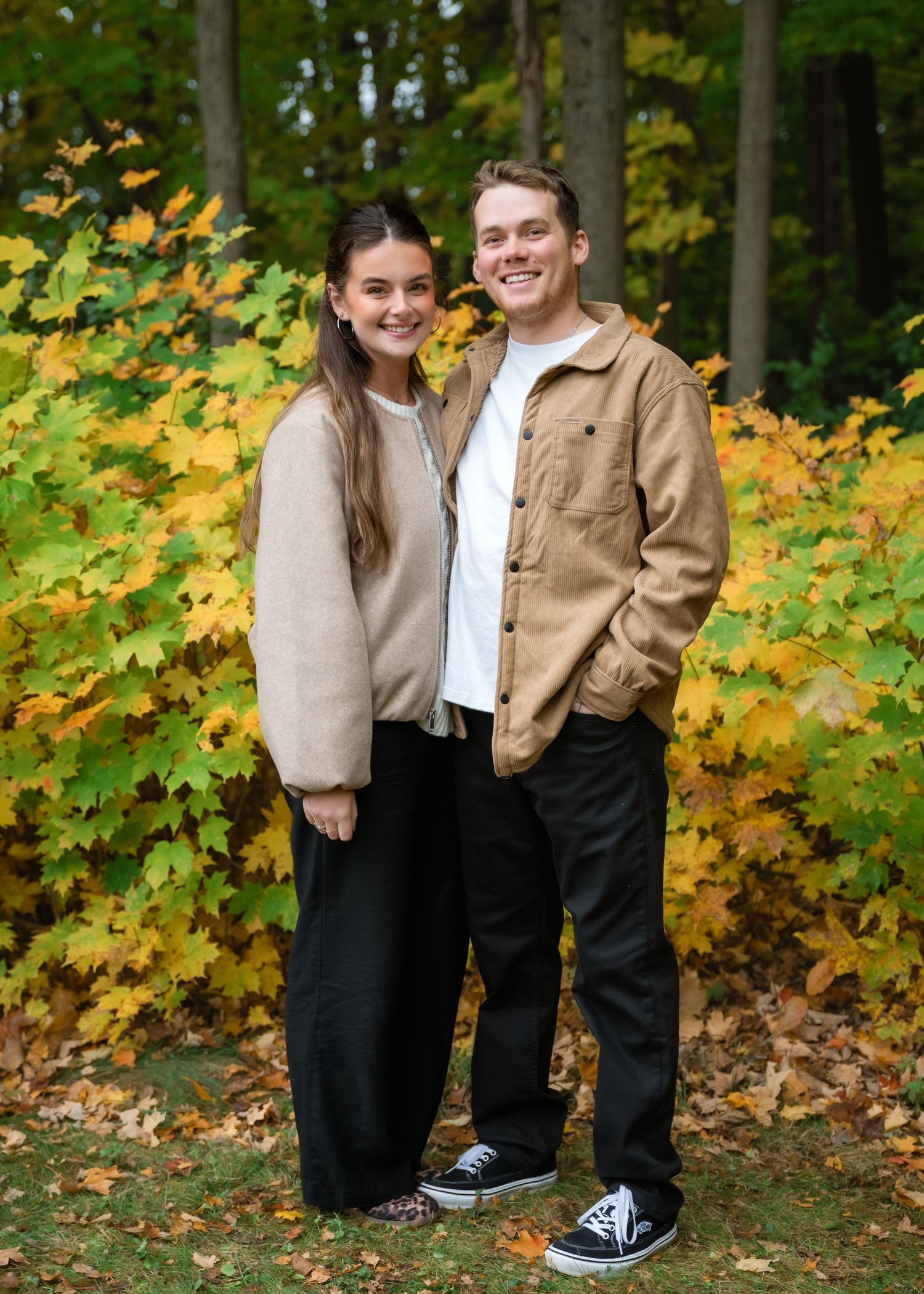 A young couple smiles together outdoors surrounded by vibrant autumn foliage in yellow and green hues.