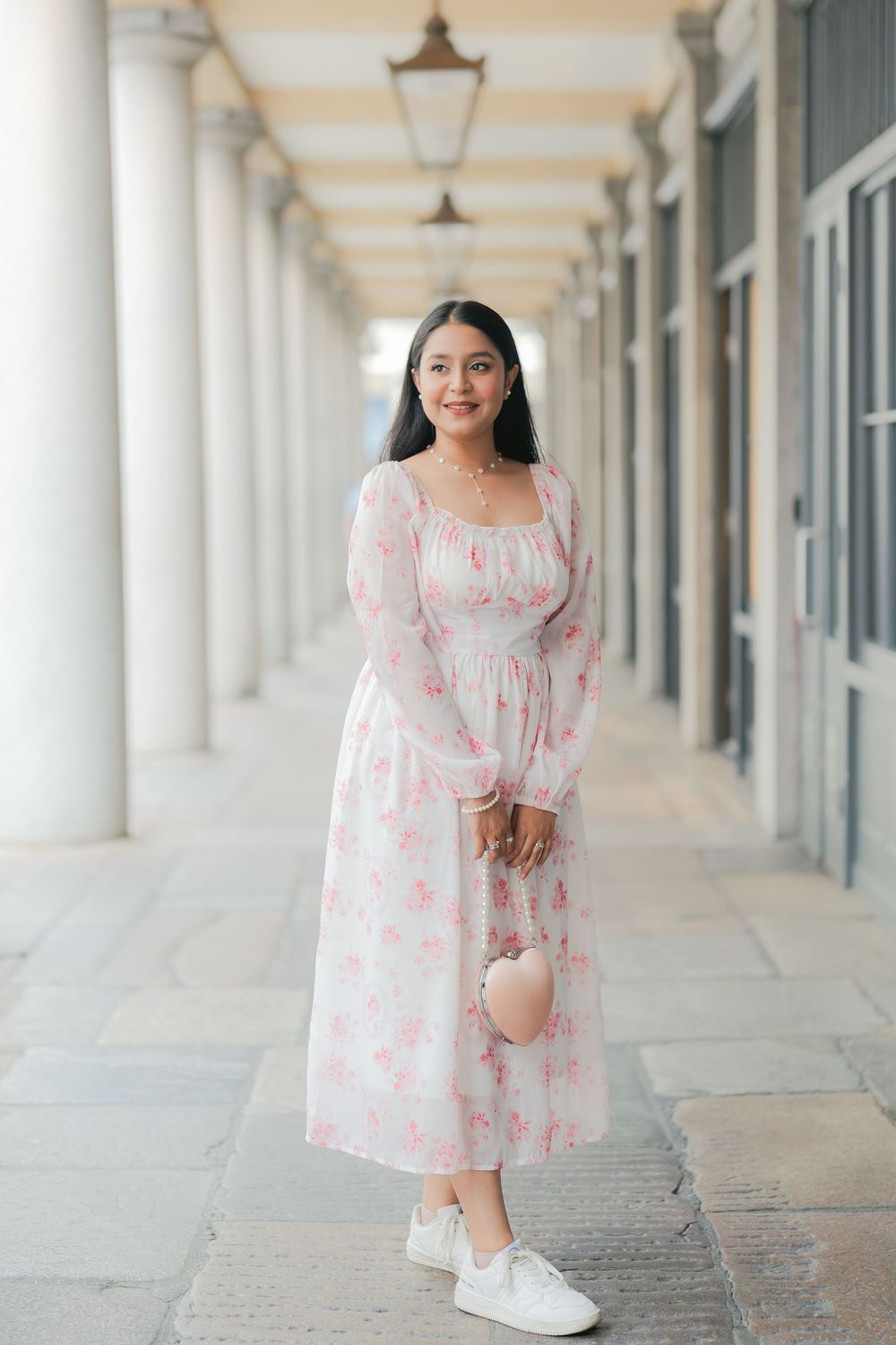 Woman in white floral midi dress with pink roses and long sleeves posing in elegant colonnade walkway.