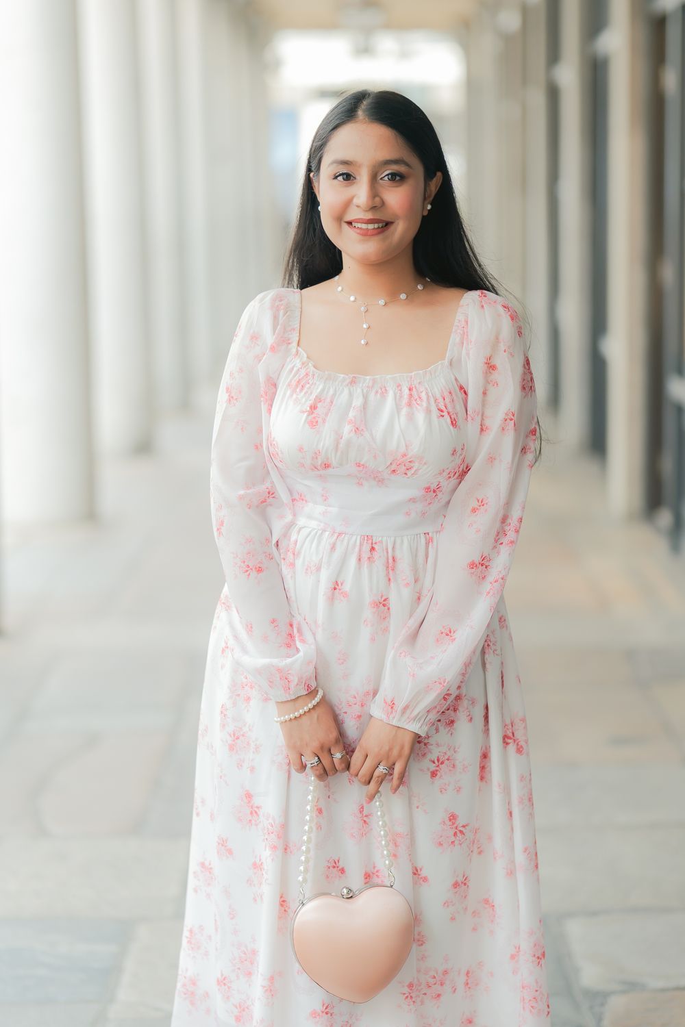 Woman in white floral dress holding heart-shaped pink purse, smiling in elegant outdoor corridor.