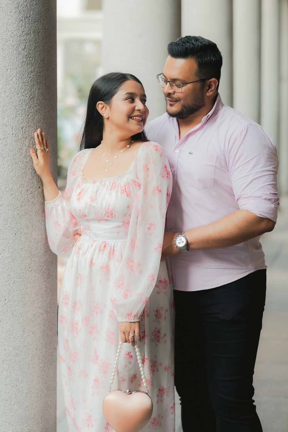 Couple posing romantically near a pillar, woman in floral dress holding heart purse, man in pink shirt wearing glasses.