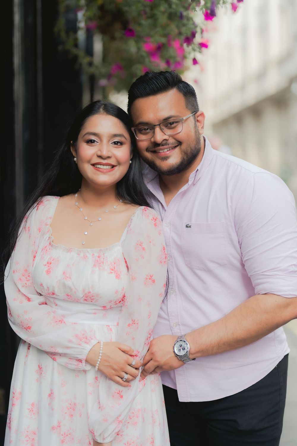 A smiling couple poses together, woman in floral white dress and man in pink shirt with glasses, near pink flowers.
