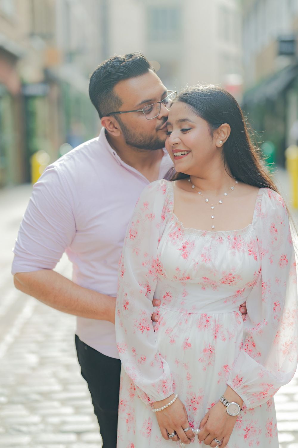 A man kisses a smiling woman in a floral dress on a cobblestone street in a romantic outdoor setting.