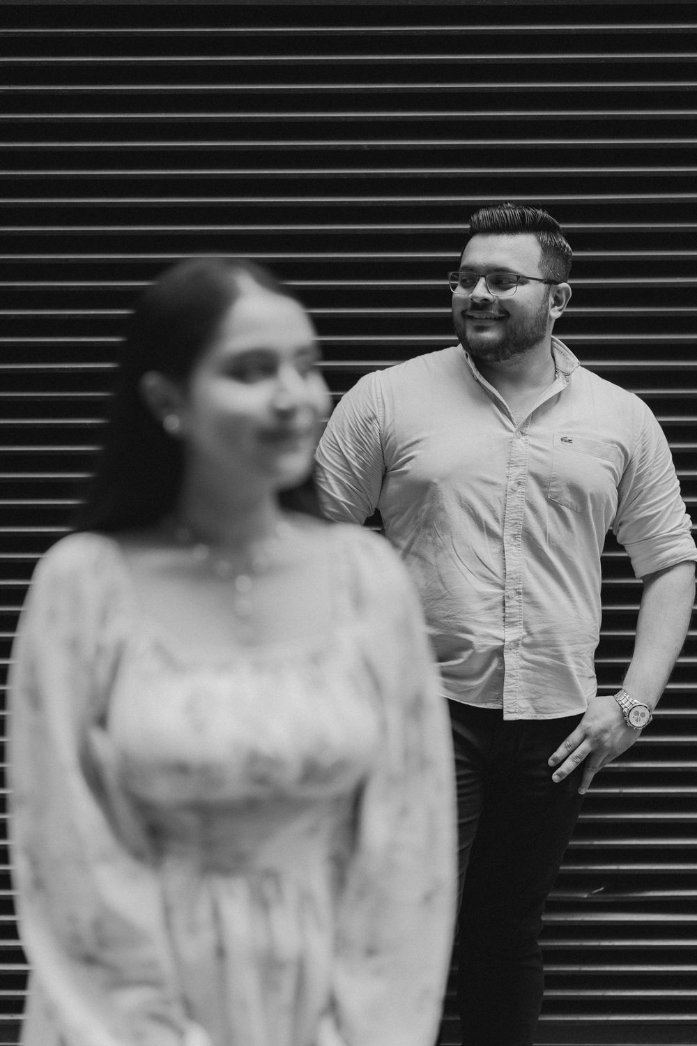 Black and white photo of a man in glasses smiling at a woman in a floral dress standing in the foreground.