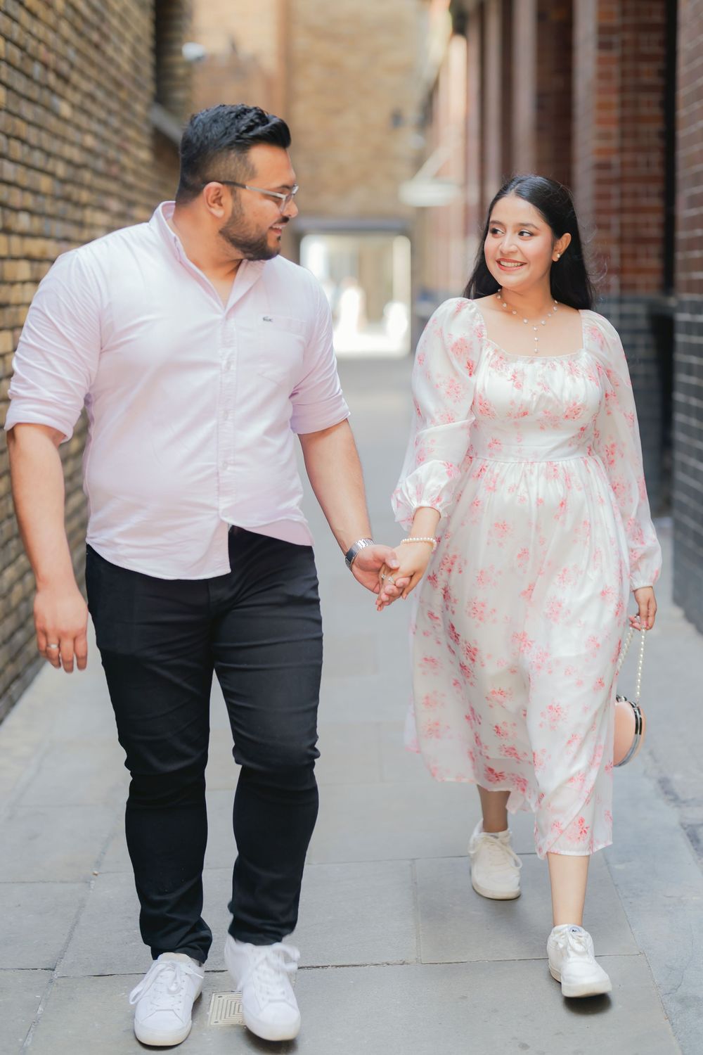 Couple holding hands walking together, man in pink shirt and black pants, woman in floral dress, urban alley backdrop.