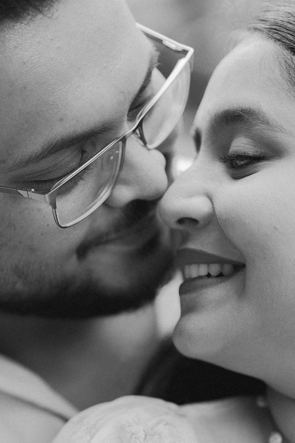 Black and white close-up of a couple sharing a tender moment, man with glasses leaning in to kiss smiling woman.