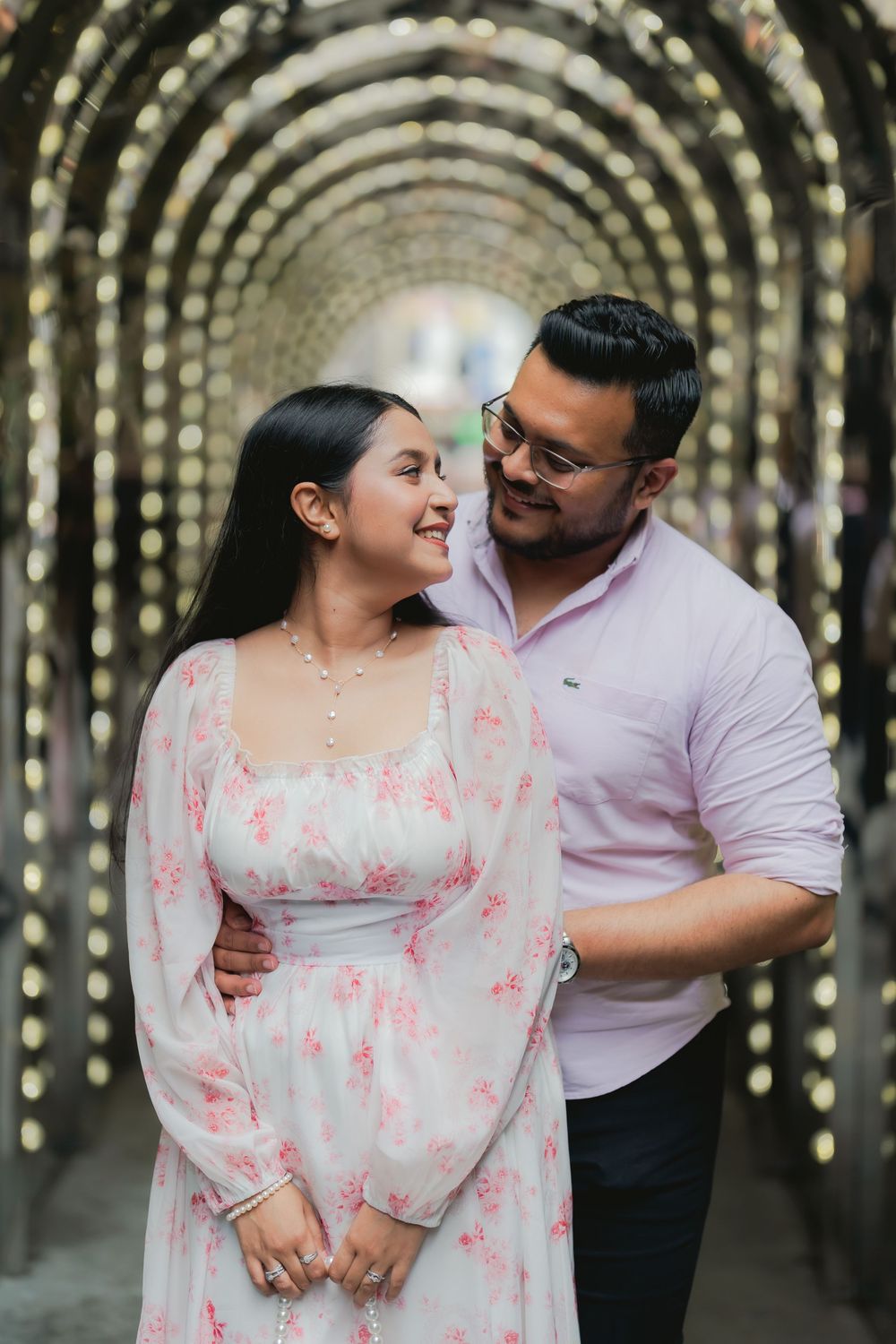 Couple smiling at each other under an illuminated archway, woman in floral dress, man in pink shirt.