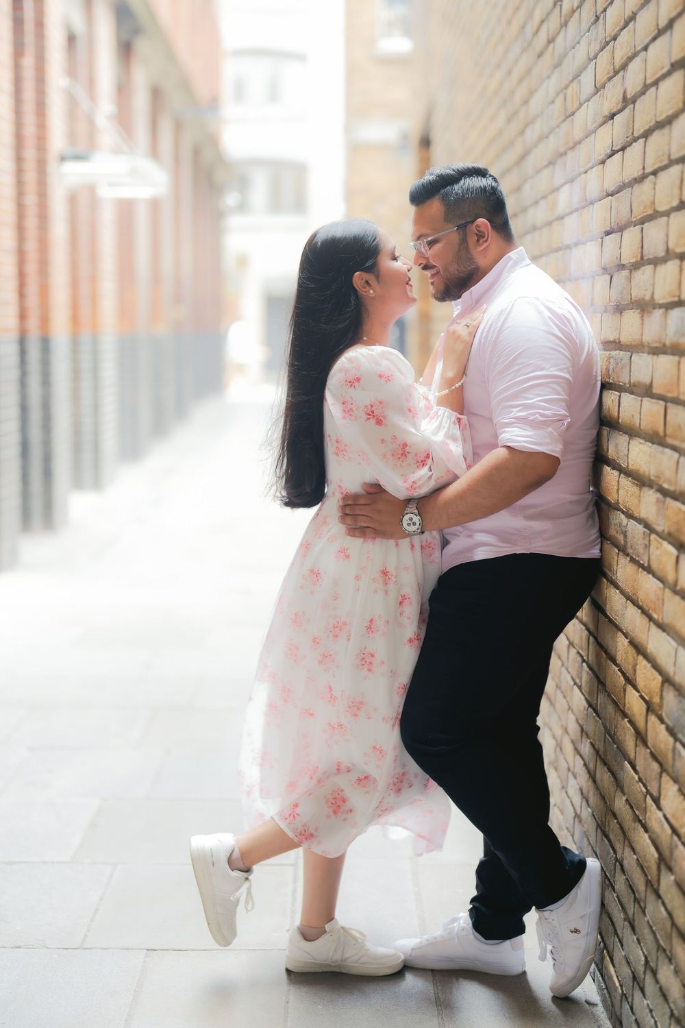 A couple shares a romantic moment leaning against a brick wall in an urban alley, woman in floral dress, man in pink shirt.