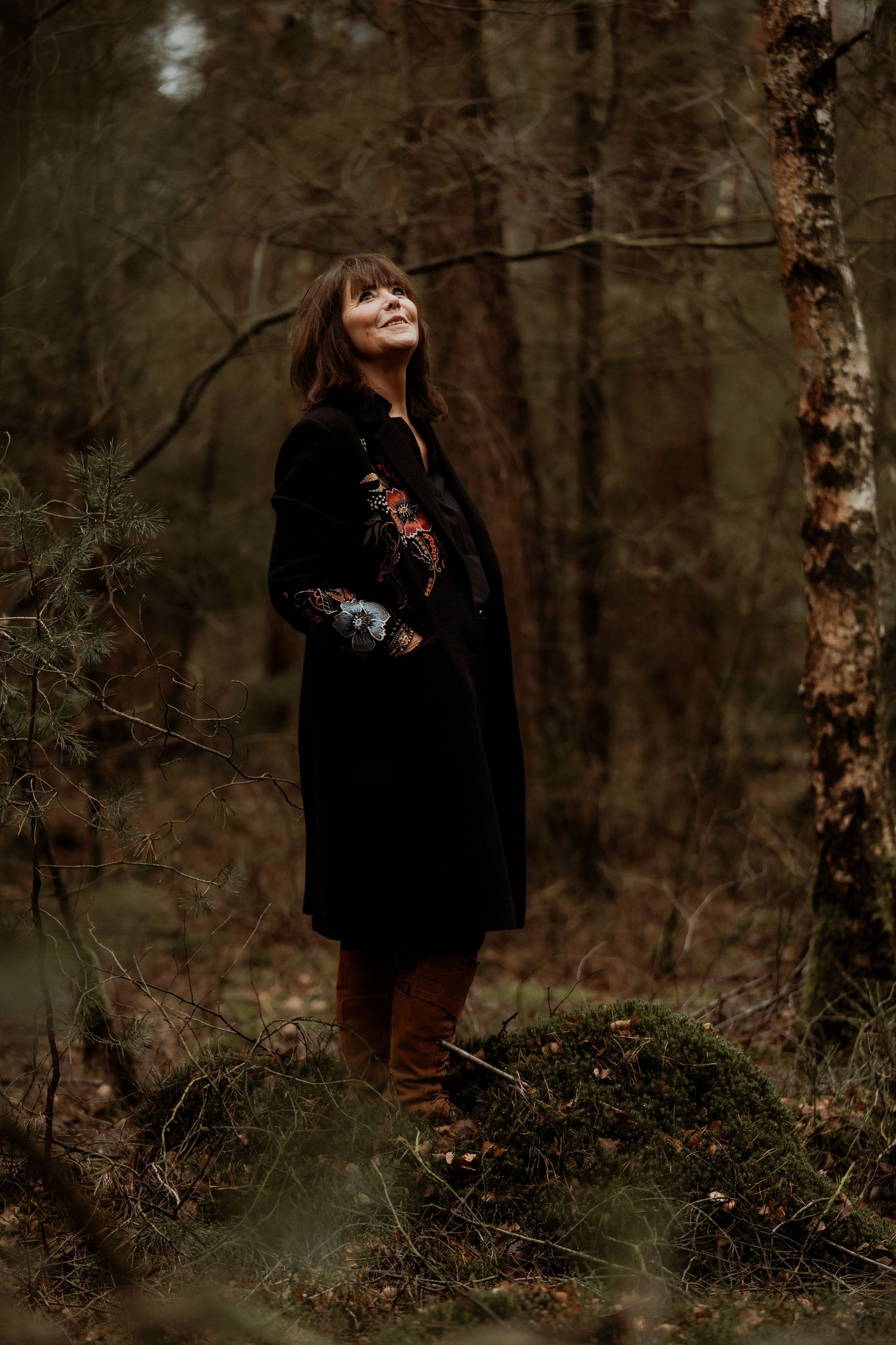 Woman in black embroidered coat standing on moss in autumn forest, gazing upward among bare trees.