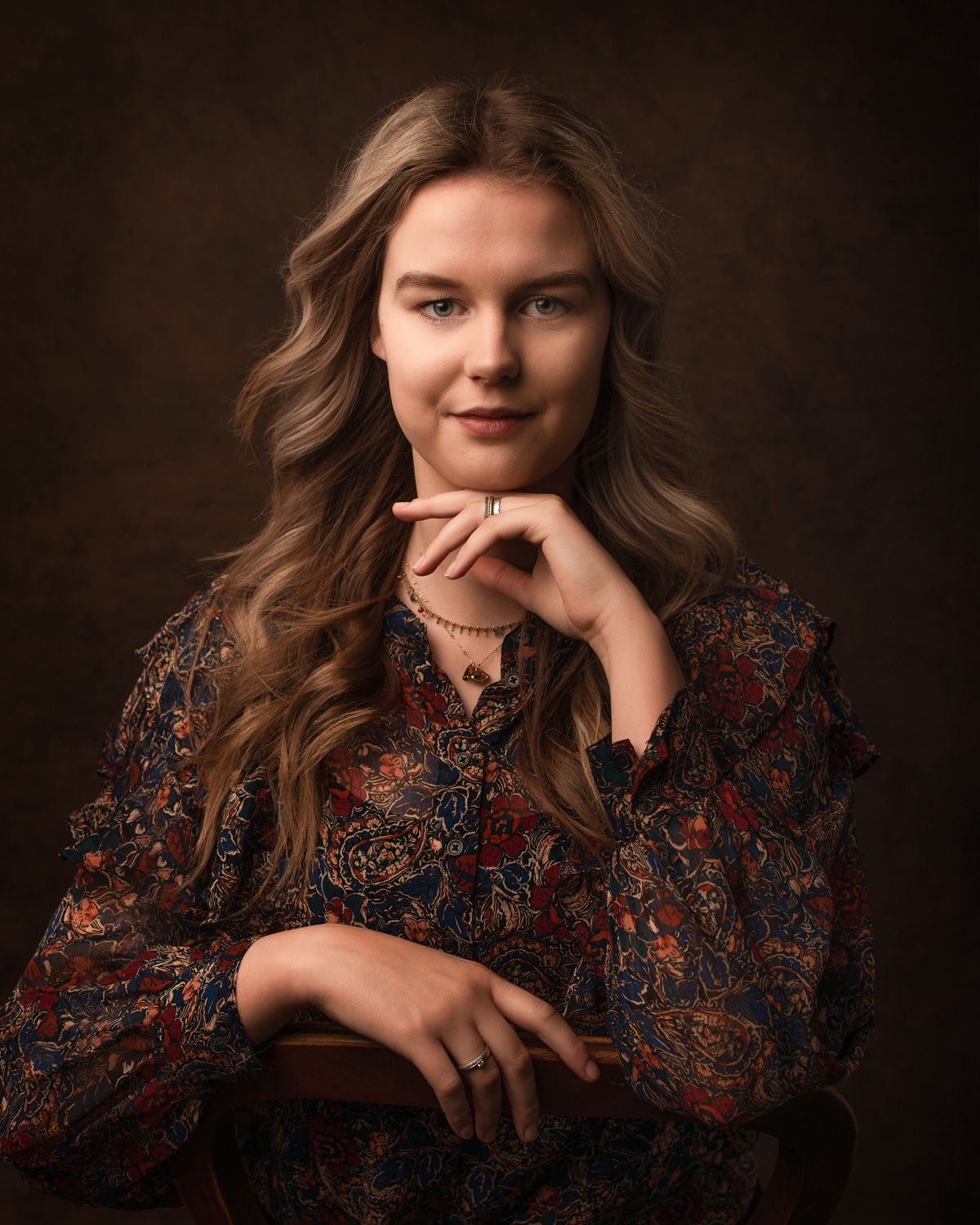 Portret in warme tinten met lichtbruin haar in een paisleyblouse met patroon tegen een donkerbruine achtergrond.