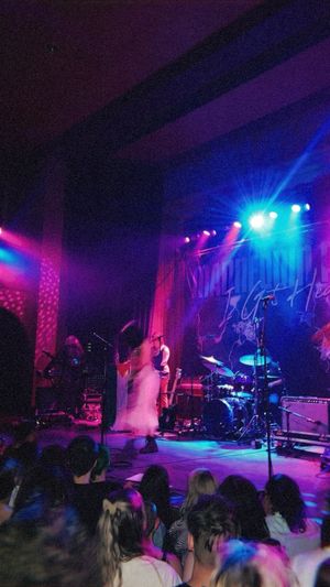 Concert performers on stage bathed in vibrant purple and blue lighting while crowd watches from below at intimate venue.