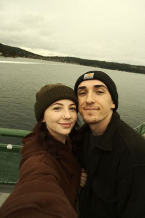 A couple wearing winter hats takes a selfie together on a ferry with water and forested coastline in the background.