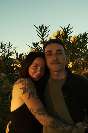 A couple sits together outdoors at sunset with trees and foliage silhouetted against a warm golden sky.