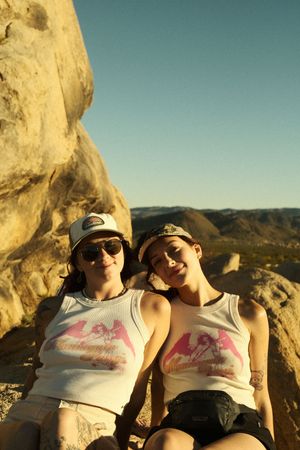 Two people relax together against a rocky backdrop in matching white tank tops at sunset.
