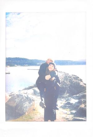 A person and child stand together on rocky coastal terrain overlooking a scenic waterway on a bright day.