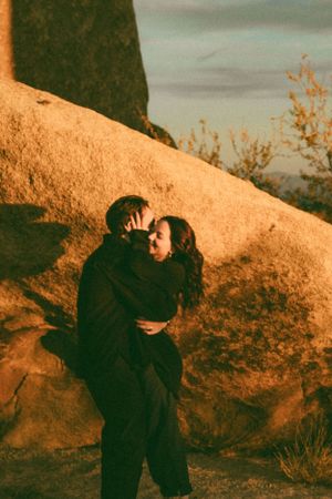 Two people embrace against a rocky desert backdrop in warm golden light at sunset.
