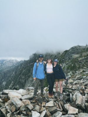 Three hikers pose together on rocky mountain terrain with misty peaks and cloudy skies in the background.