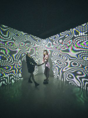 Two figures dance in a dark room with swirling psychedelic light projections creating mesmerizing patterns on the walls.