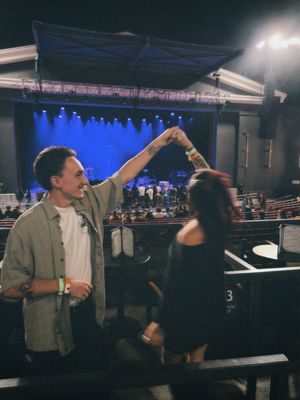 People interact and gesture on a dimly lit stage with blue lighting and theater seating visible in the background.