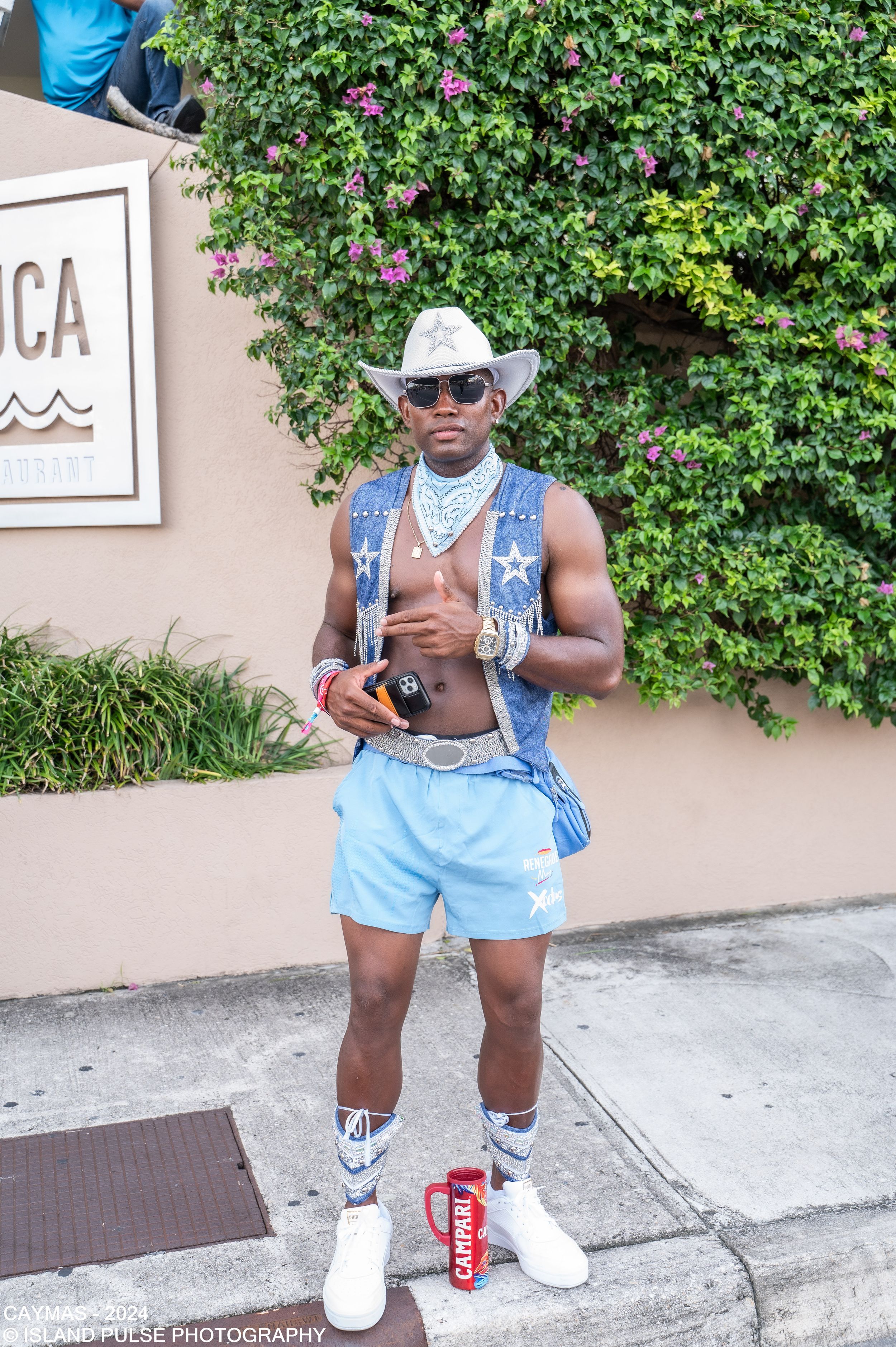 Man in blue cowboy outfit with hat, sunglasses, denim vest, and white boots posing outdoors at a festival.